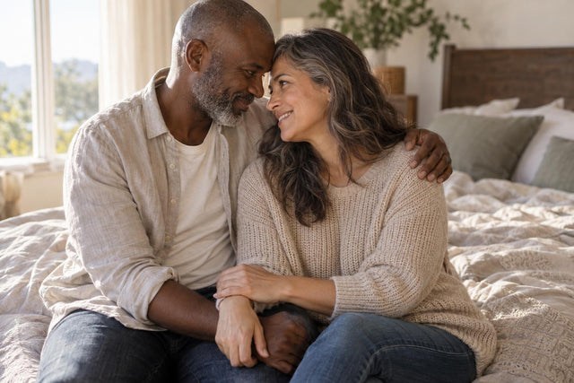 Couple in their forties sitting close on a bed, laughing together in soft morning light
