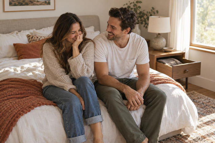 Couple in casual clothes laughing together on the edge of a bed in soft morning light
