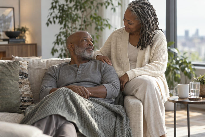 Man resting on a sofa under a blanket while his partner rests a hand on his shoulder